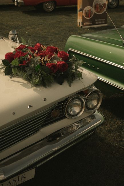 A floral arrangement featuring a bouquet of fresh red roses, white berries, and lush green foliage, resting on the hood of a vintage silver car. The bouquet is wrapped with a subtle, transparent wrapp