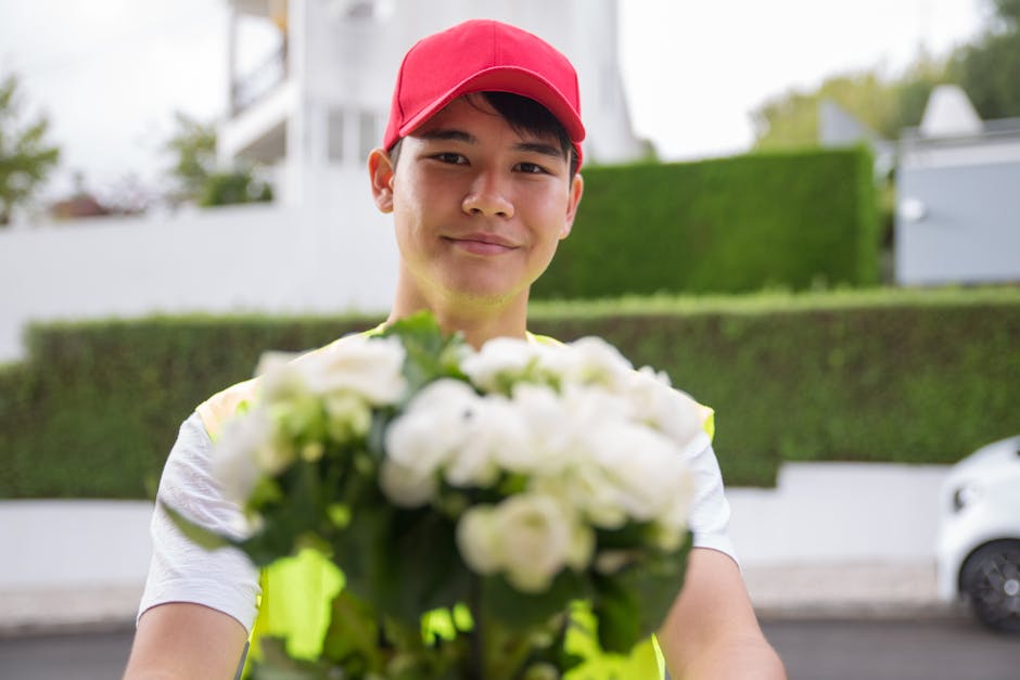 A young delivery person wearing a red cap and a high-visibility vest is outdoors, holding a bouquet of fresh, white roses with green foliage. The bouquet is arranged in a natural, rounded style and wr