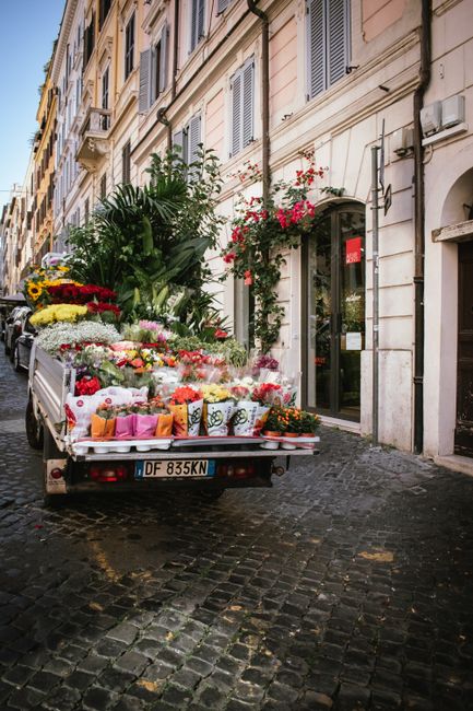 A white delivery truck parked on a cobblestone street in front of a building façade with beige and cream-colored walls, featuring decorative moldings and window shutters. The truck is loaded with a v