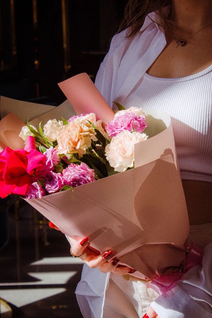 A woman holding a bouquet of fresh flowers wrapped in beige paper, featuring pink, white, and light peach blooms, including carnations and roses, with green foliage accents. She is wearing a white top