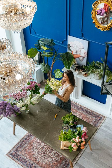A woman in a beige top and patterned skirt is arranging a bouquet of fresh flowers on a grey table inside a bright, stylish room with bold blue walls. The floral arrangement includes white, green, and