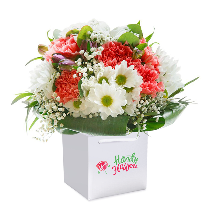 A beautifully realised bouquet rises from the heart of a sleek white gift bag, its full, rounded form brimming with the life and colour that characterise Old Ford. The centre is awash with ruffled coral and peach carnations, their generous, layered petals offering a tactile softness, while strategic dashes of vibrant red deepen the visual richness. Dotted throughout, clusters of white daisy-like flowers with radiant lime-green centres evoke the innocence and energy of wild blooms along the Regent's Canal path. Baby's breath, in delicate white sprays, brings lightness and a sense of air, while well-placed purple-tinged buds-subtly closed-introduce intrigue and ever-present potential. Dense, broad green foliage and glossy leaves wrap around the base, creating a botanical collar that grounds the whole with Old Ford's trademark greenery. This bouquet is styled for both distinction and accessibility, the clean white bag with its sturdy ribbon handles conveying both elegance and practical delivery-a familiar sight for those living in warehouse flats or historic terraces off Tredegar Road. The professional composition and pure white backdrop highlight every detail, from petal veining to the interplay of colour, ensuring the arrangement stands out as a ready-to-gift treasure. This arrangement is perfect for marking a milestone, brightening a flat on a rainy East London afternoon, or sharing joy with loved ones nearby-celebrating the freshness and artistry characteristic of local Old Ford florists.