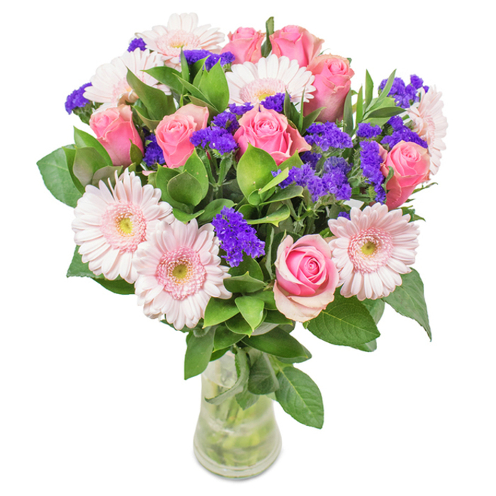 An expertly constructed floral centerpiece rests in a clear glass vase, poised against a spotless white backdrop-its colours and elegance reminiscent of blooms found at Old Ford's historic Roman Road markets. The arrangement features a central grouping of seven to nine medium-to-soft pink roses, each gracefully spiraling from bud to half-open bloom. These are partnered by five to seven pale pink gerbera daisies, identifiable by their prominent, round heads and lighter, almost silvery centers. Scattered among the larger blossoms are eye-catching clusters of deep purple statice, which lend striking texture and definition. Rich, leafy green foliage provides a sense of abundance, echoing the vibrancy of local East London parks. The vase, with stems immersed in clear water, underscores the bouquet's freshness and authenticity. Designed for any occasion-be it birthdays, anniversaries, Mother's Day, or simple home adornment-this arrangement of roses, gerberas, and statice flowers communicates joy, celebration, and attentive care, promising an Old Ford delivery imbued with thoughtfulness.